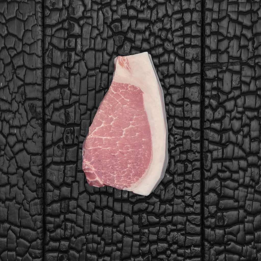 Rohes Duroc-Karree-Steak, hellrosa Fleischfarbe mit feiner Marmorierung und gleichmäßigem, breitem Fettrand, kompakte, leicht gebogene Form, mittig auf dunkler, verkohlter Holzoberfläche.