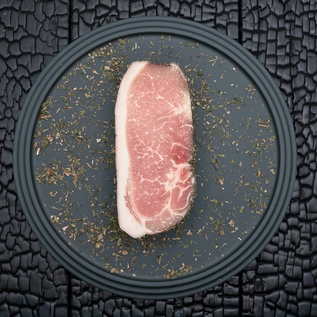 Rohes Duroc-Karree-Steak, hellrosa Fleischfarbe mit feiner Marmorierung und gleichmäßigem Fettrand, mit getrockneten Alpenkräutern umgeben, mittig auf einem dunklen Teller platziert, vor dunkler, verkohlter Holzoberfläche.