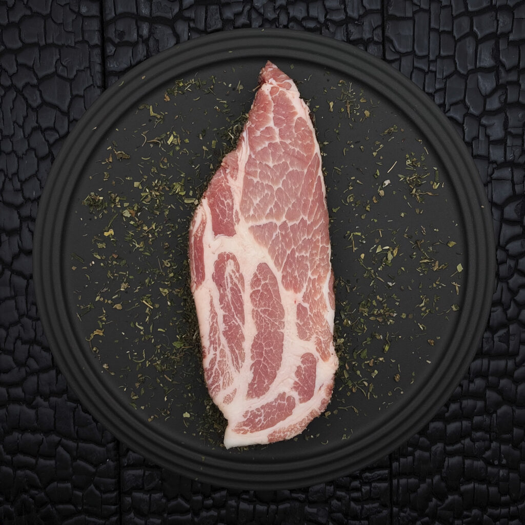 Rohes Stück Duroc Schopf, gereift mit Alpenkräutern, liegt zentral auf einem runden schwarzen Teller. Das rosarote Fleisch zeigt eine feine weiße Fettmarmorierung mit deutlich abgegrenzten Muskelpartien. Rund um das Fleisch sowie auf dem Teller sind getrocknete Kräuter verstreut. Der Teller liegt auf einer schwarz verkohlten Holzoberfläche mit stark strukturierter, rissiger Maserung.