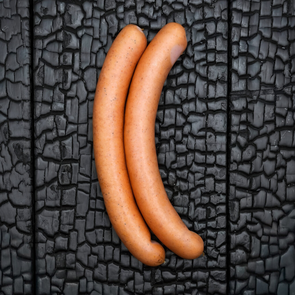 Zwei Frankfurter Würstel, hellbraune bis goldene Oberfläche, glatt und leicht glänzend, gebogene Form, parallel nebeneinander liegend, mittig auf dunkler, verkohlter Holzoberfläche.