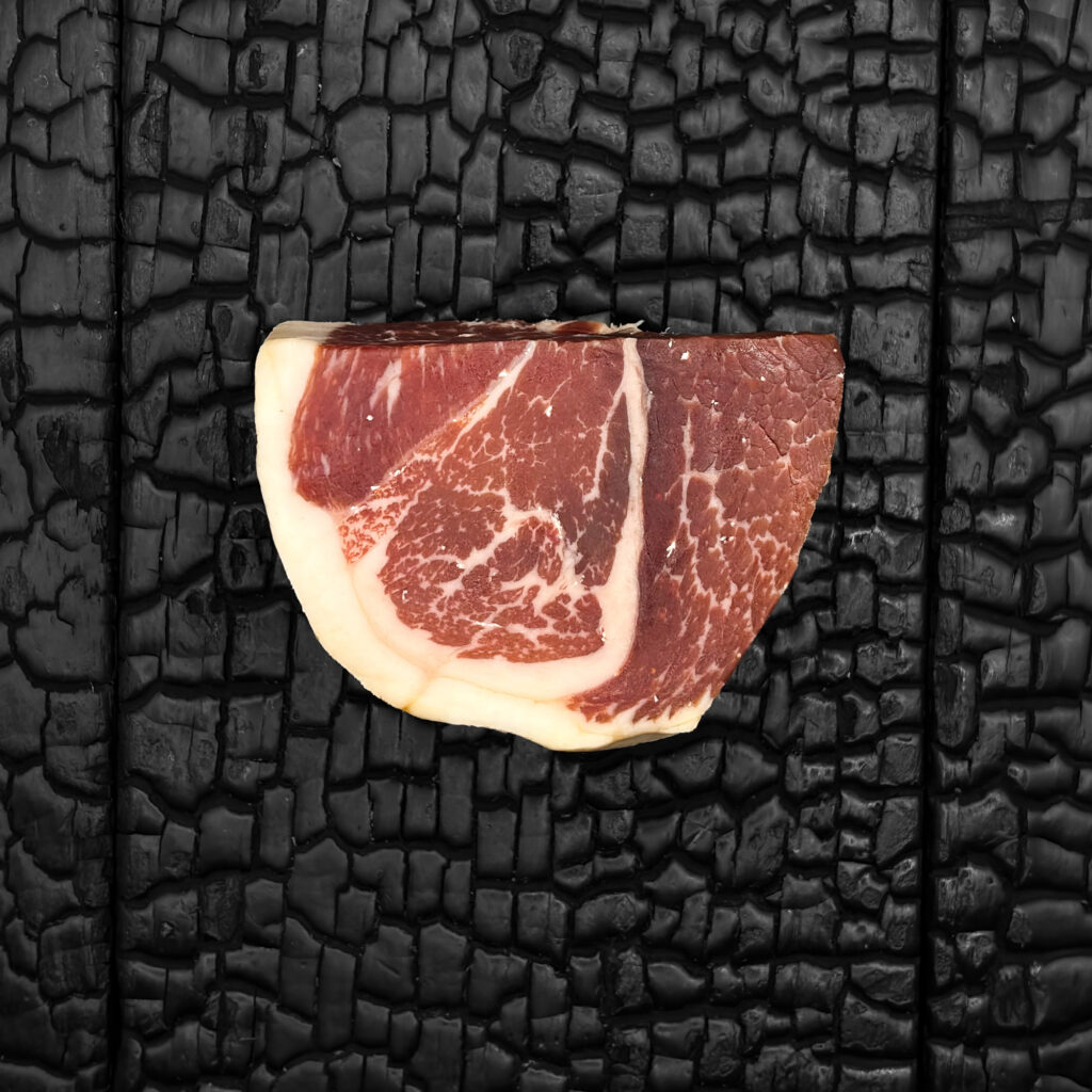 Jamón-Stück mit dunkelroter Fleischfarbe und feiner, gleichmäßiger Marmorierung, umrandet von einem hellen Fettdeckel. Kompakte Form, mittig auf einer dunklen, verkohlten Holzoberfläche platziert.