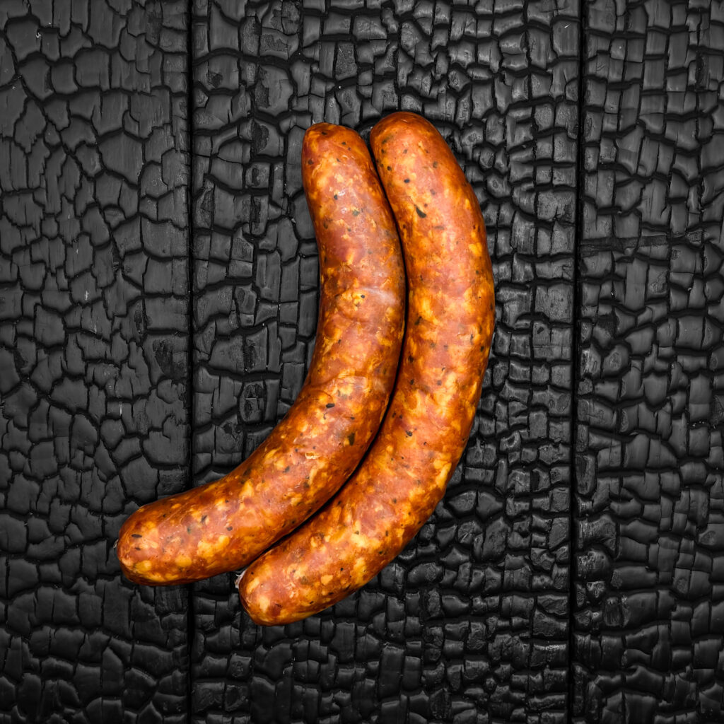 Zwei Chili Käsekrainer, hellbraune Oberfläche mit sichtbaren Käse- und Gewürzeinschlüssen, leicht glänzend, gebogene Form, eng nebeneinander liegend, mittig auf dunkler, verkohlter Holzoberfläche.