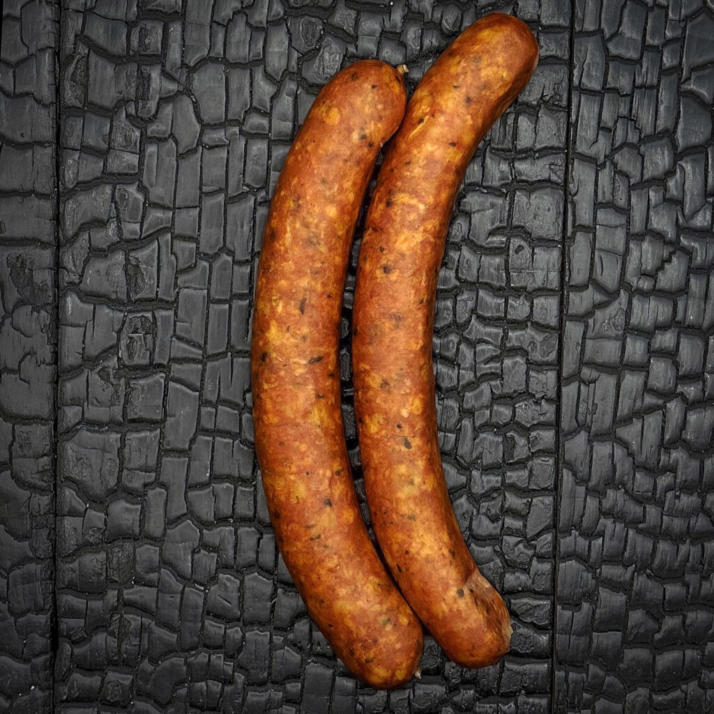 Zwei gebogene rohe Käswürstel liegen parallel auf einer schwarz verkohlten Holzoberfläche mit stark strukturierter, rissiger Maserung. Die Würstel zeigen eine rotbraune Oberfläche mit sichtbaren Gewürz- und Käsestücken. Der starke Kontrast zum dunklen Hintergrund hebt Form, Struktur und die handwerkliche Verarbeitung der Würstel hervor.