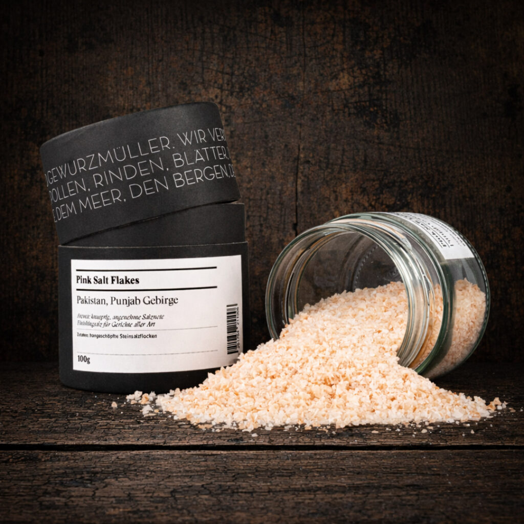 Ein schwarzer Gewürzbehälter mit leicht geöffnetem Deckel steht links im Bild. Auf dem Etikett ist „Pink Salt Flakes“ sowie „Pakistan, Punjab Gebirge“ zu lesen. Rechts daneben liegt ein geöffnetes Glas auf der Seite, aus dem rosafarbene, grobe Salzflocken herausgeschüttet wurden und einen kleinen Haufen im Vordergrund bilden. Die Szene ist mittig auf einer dunklen, rustikalen Holzoberfläche arrangiert.