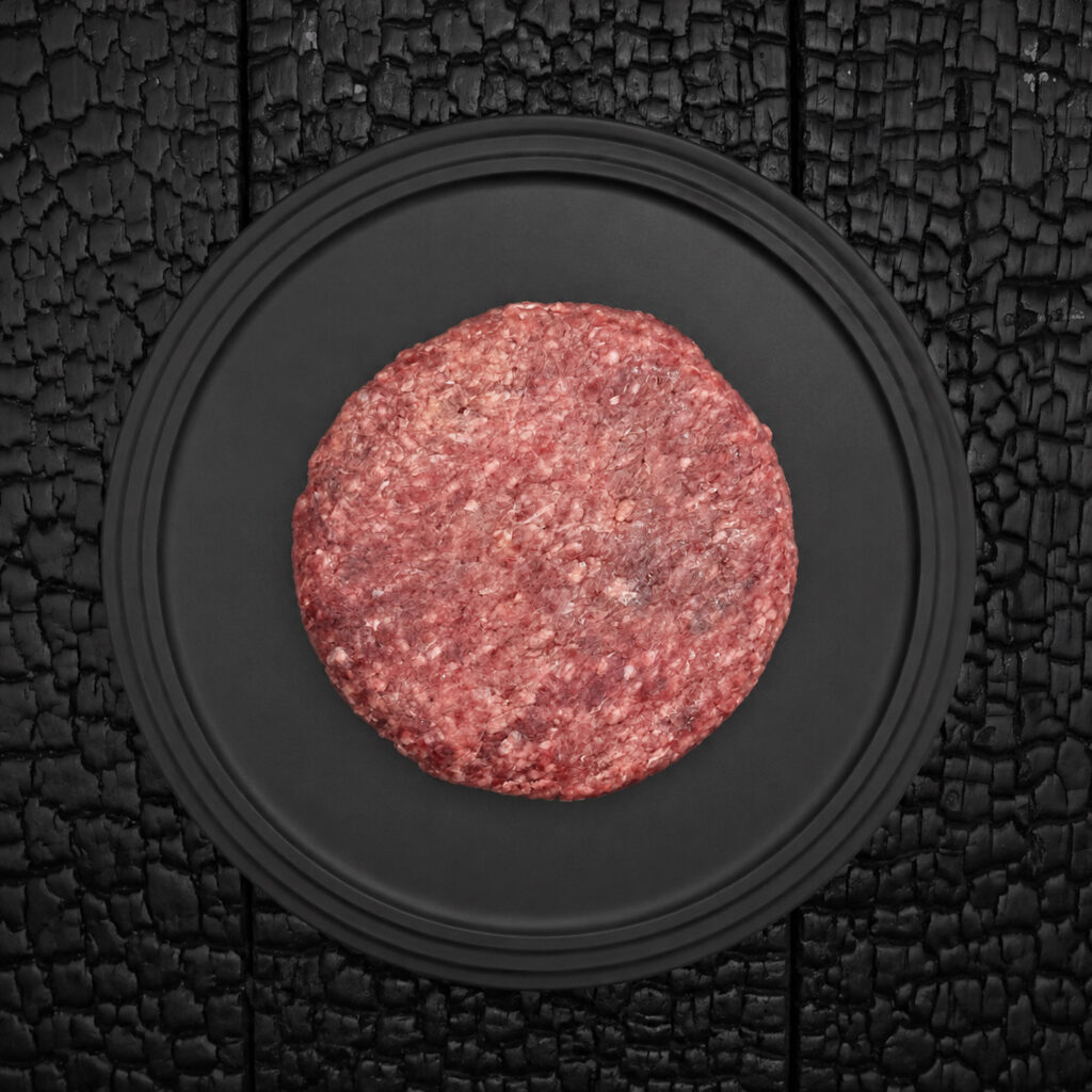Ein rohes Burger Patty (dry aged Wagyu Rindfleisch) liegt zentral auf einem runden, schwarzen Teller. Das Fleisch zeigt eine grob gewolfte Struktur mit sichtbaren hellen Fettanteilen und einer kräftig roten bis rosafarbenen Fleischfarbe. Die runde Form des Patties wirkt gleichmäßig gepresst und handwerklich hergestellt. Der Teller befindet sich auf einer schwarz verkohlten Holzoberfläche mit stark strukturierter, rissiger Maserung. Der dunkle Hintergrund hebt die Farbe und Struktur des Fleisches deutlich hervor. Die Aufnahme erfolgt aus direkter Draufsicht und wirkt reduziert, symmetrisch und grafisch. Der Fokus liegt auf der Fleischstruktur, dem Fettanteil und der hochwertigen Präsentation des Burger Patties. Die Darstellung vermittelt handwerkliche Fleischverarbeitung, hochwertige Fleischqualität und eine stilvolle Produktinszenierung.