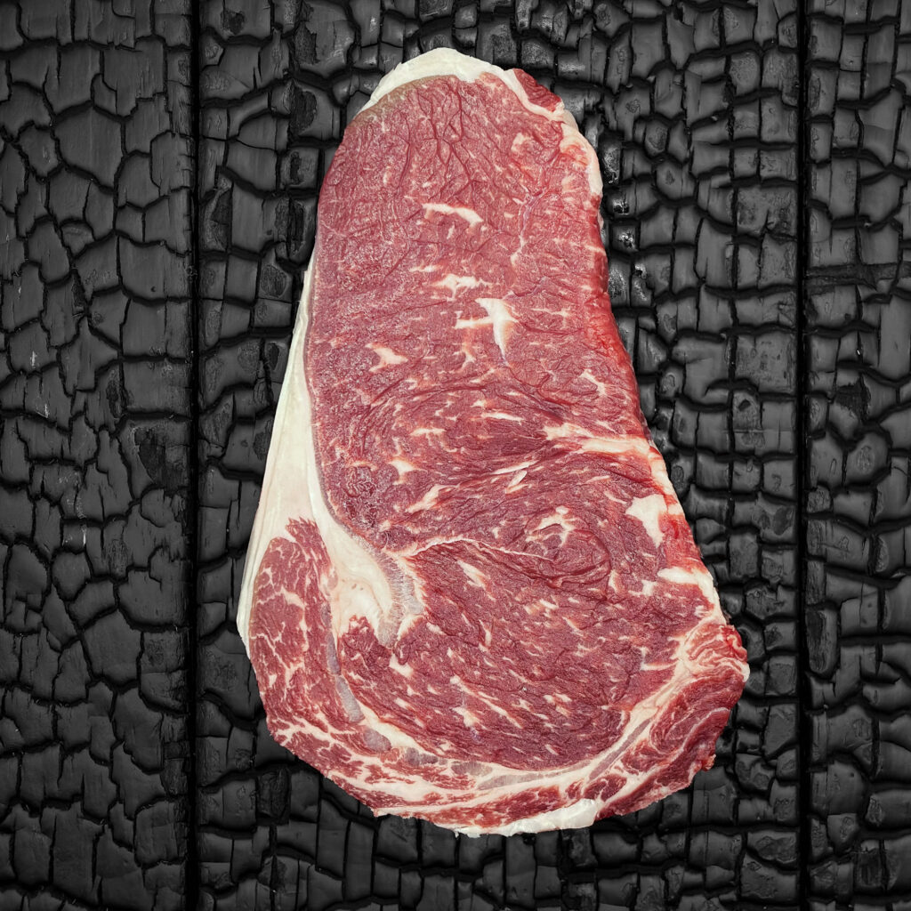 Rohes Entrecôte-Steak vom Rind mit feiner Marmorierung, rote Farbe, Fettauge und Fettrand sichtbar, mittig auf dunkler, verkohlter Holzoberfläche.
