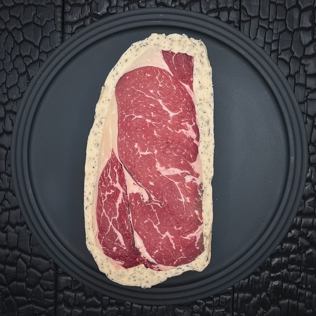 Ein rohes Entrecote Steak liegt zentral auf einem runden schwarzen Teller. Das dunkelrote Fleisch zeigt eine feine weiße Fettmarmorierung und ist von einem hellen Kräuterbuttermantel mit sichtbaren Kräuterstücken umgeben. Der Teller liegt auf einer schwarz verkohlten Holzoberfläche mit stark strukturierter, rissiger Maserung.
