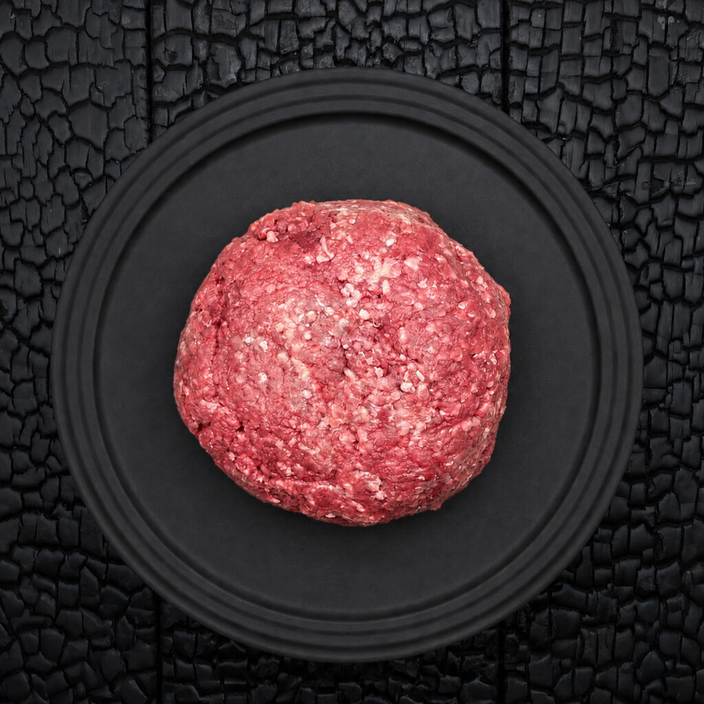 Ein großer Ballen rohes Faschiertes beziehungsweise Hackfleisch vom Rind (dry aged) liegt zentral auf einem runden, schwarzen Teller. Das Fleisch zeigt eine grob gewolfte Struktur mit sichtbaren hellen Fettanteilen und einer kräftig roten bis rosafarbenen Farbe. Die Oberfläche wirkt locker geformt und handwerklich verarbeitet. Der schwarze Teller liegt auf einer schwarz verkohlten Holzoberfläche mit stark strukturierter, rissiger Maserung. Der dunkle Hintergrund hebt die Farbe und Struktur des Fleisches deutlich hervor und erzeugt einen starken Kontrast. Die Aufnahme erfolgt aus direkter Draufsicht und wirkt reduziert, symmetrisch und grafisch. Der Fokus liegt auf der Fleischstruktur, dem Fettanteil und der hochwertigen Präsentation des frischen Hackfleischs. Die Darstellung vermittelt handwerkliche Fleischverarbeitung, hochwertige Fleischqualität und eine stilvolle Produktinszenierung.