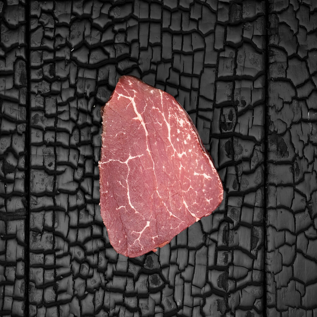 Rohes Filetsteak vom Rind mit feiner Marmorierung, rote Fleischfarbe, einzelnes Stück auf dunkler, verkohlter Holzoberfläche.