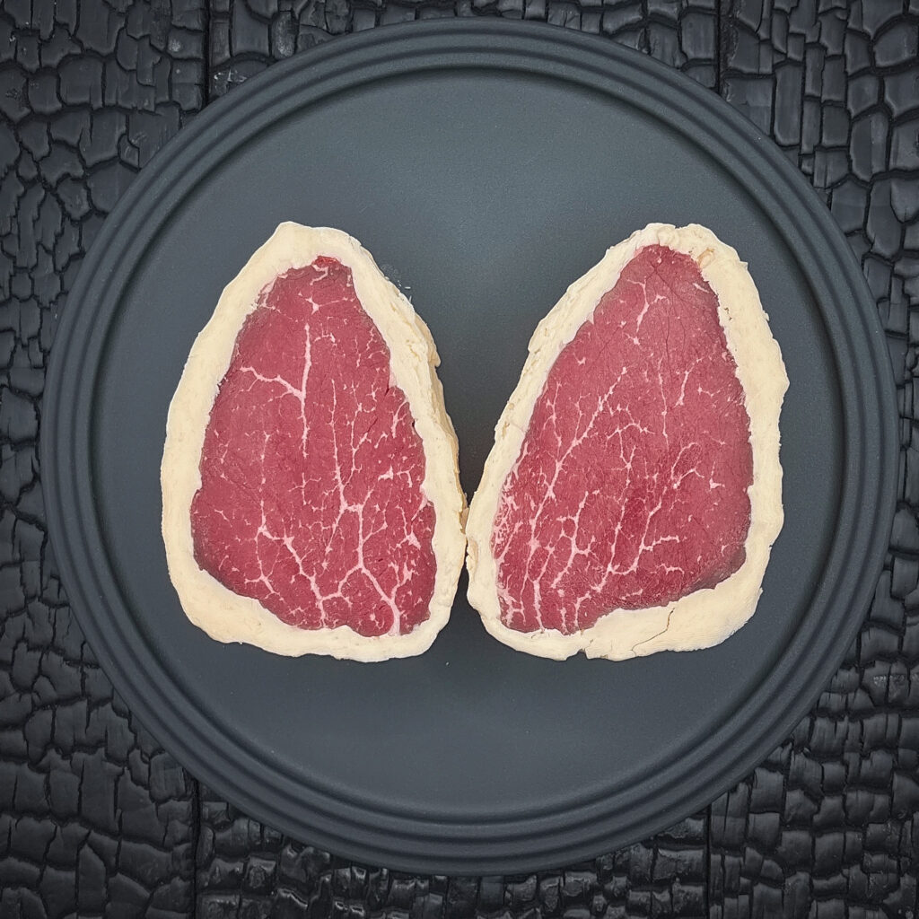 Zwei rohe Rinderfiletsteaks liegen spiegelbildlich nebeneinander auf einem runden schwarzen Teller. Das dunkelrote Fleisch zeigt eine feine Fettmarmorierung und ist vollständig von einem hellen Buttermantel umgeben. Der Teller liegt auf einer schwarz verkohlten Holzoberfläche mit stark strukturierter, rissiger Maserung.