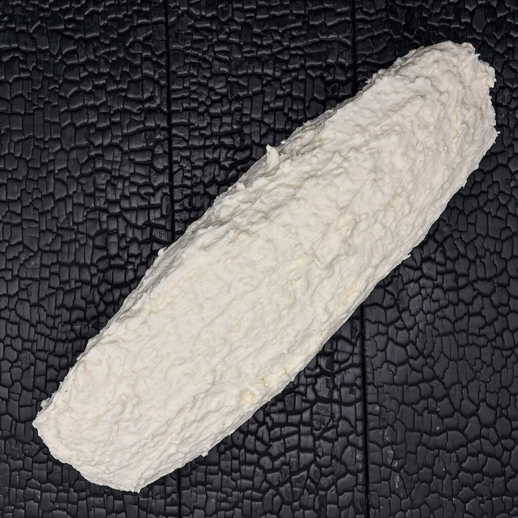 Ein längliches Stück Rinderfilet liegt diagonal auf einer schwarz verkohlten Holzoberfläche mit stark strukturierter, rissiger Maserung. Das Fleischstück ist vollständig von einer dicken, weißen Butterummantelung bedeckt. Die Oberfläche wirkt unregelmäßig, cremig und handwerklich aufgetragen. Durch die helle Ummantelung hebt sich das Fleischstück deutlich vom dunklen Hintergrund ab. Die Aufnahme erfolgt aus direkter Draufsicht und wirkt reduziert und grafisch. Der Fokus liegt auf der Veredelung, der Struktur der Butterummantelung und der kontrastreichen Präsentation. Die Darstellung vermittelt handwerkliche Fleischreifung und besondere Veredelungstechniken.