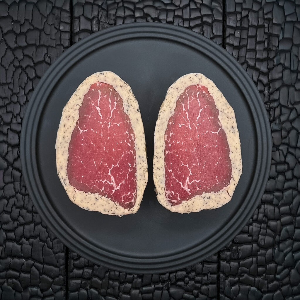Zwei rohe Rinderfiletsteaks liegen nebeneinander auf einem runden, schwarzen Teller. Die Steaks sind spiegelbildlich angeordnet und zeigen eine dunkelrote Fleischfarbe mit feiner, gleichmäßiger Marmorierung. Beide Stücke sind von einem Kräuterbuttermantel umgeben, in dem kleine dunkle Kräuterstücke sichtbar sind. Der Teller liegt auf einer schwarz verkohlten Holzoberfläche mit stark strukturierter, rissiger Maserung. Die dunkle Umgebung hebt die Butterummantelung und die Fleischstruktur deutlich hervor. Die Aufnahme erfolgt aus direkter Draufsicht und wirkt reduziert und symmetrisch. Der Fokus liegt auf der Veredelung, der Fleischqualität und dem Kontrast zwischen dunklem Hintergrund und hellem Buttermantel. Die Darstellung vermittelt hochwertige Fleischreifung und handwerkliche Veredelung.