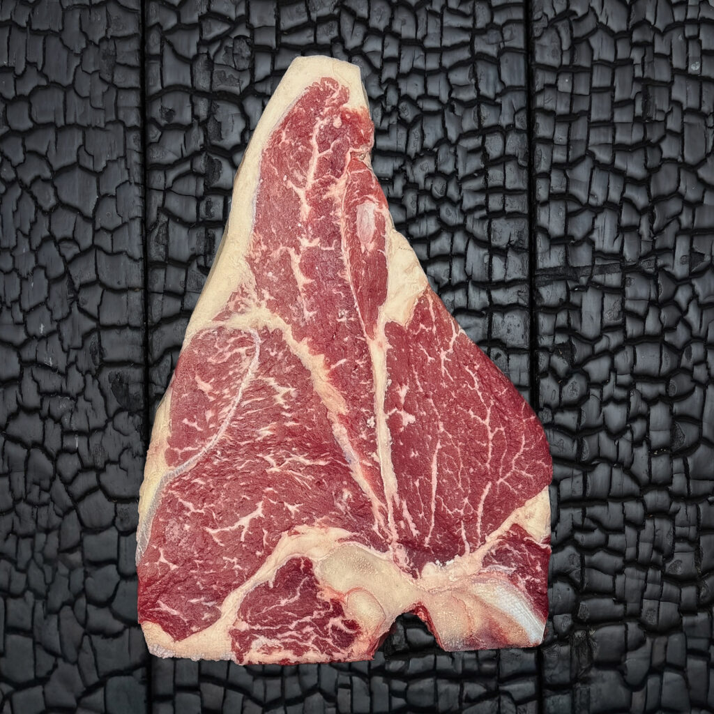 Rohes Porterhouse-Steak vom Rind mit T-förmigem Knochen, fein marmoriert, Filet- und Beiriedanteil, rote Farbe, Fettrand sichtbar, mittig auf dunkler, verkohlter Holzoberfläche.