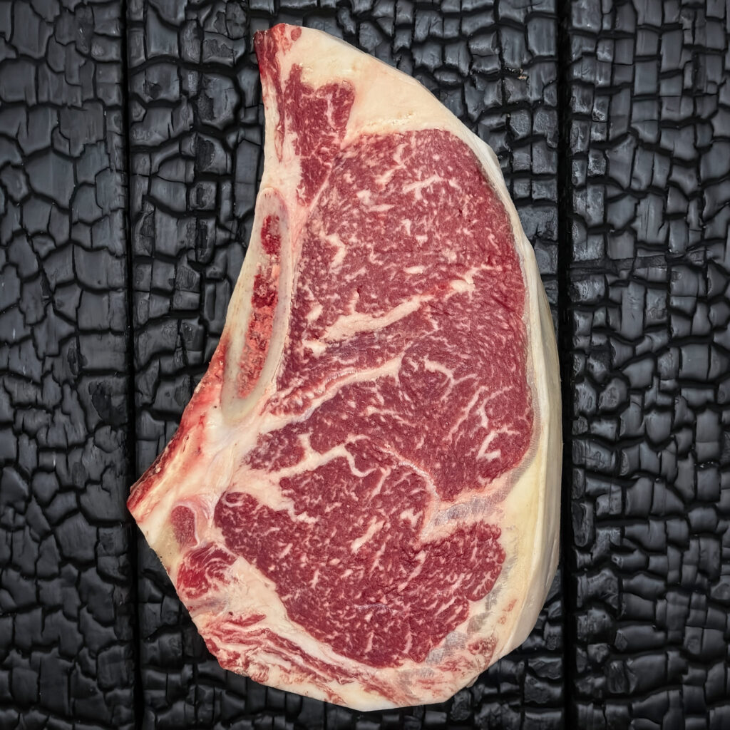 Rohes Club Steak mit Knochen vom Rind, gut marmoriert, rote Fleischfarbe mit hellem Fettrand, mittig auf dunkler, verkohlter Holzoberfläche.