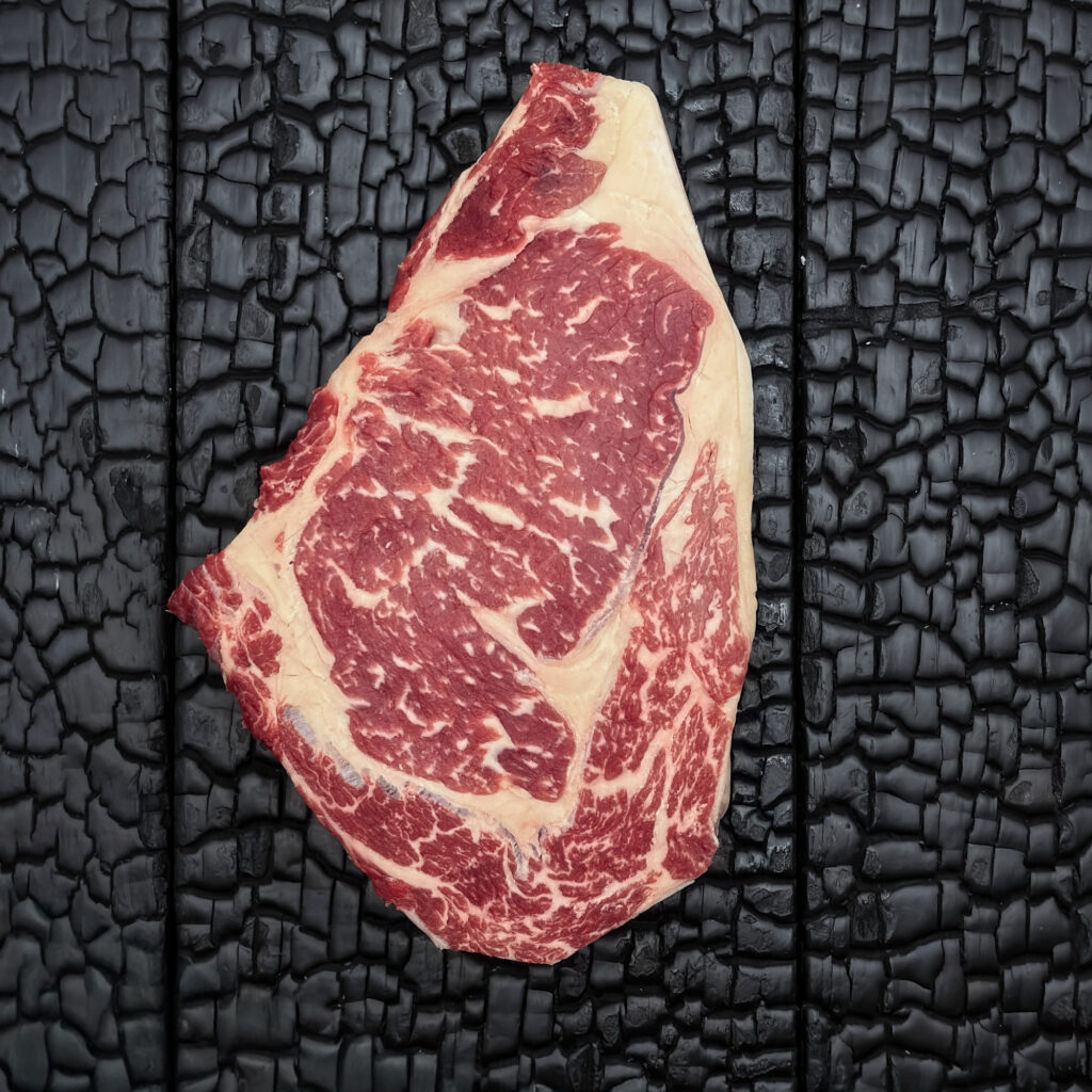 Rohes Entrecôte-Steak vom Rind mit guter Marmorierung, rote Farbe, Fettauge und Fettrand sichtbar, mittig auf dunkler, verkohlter Holzoberfläche.