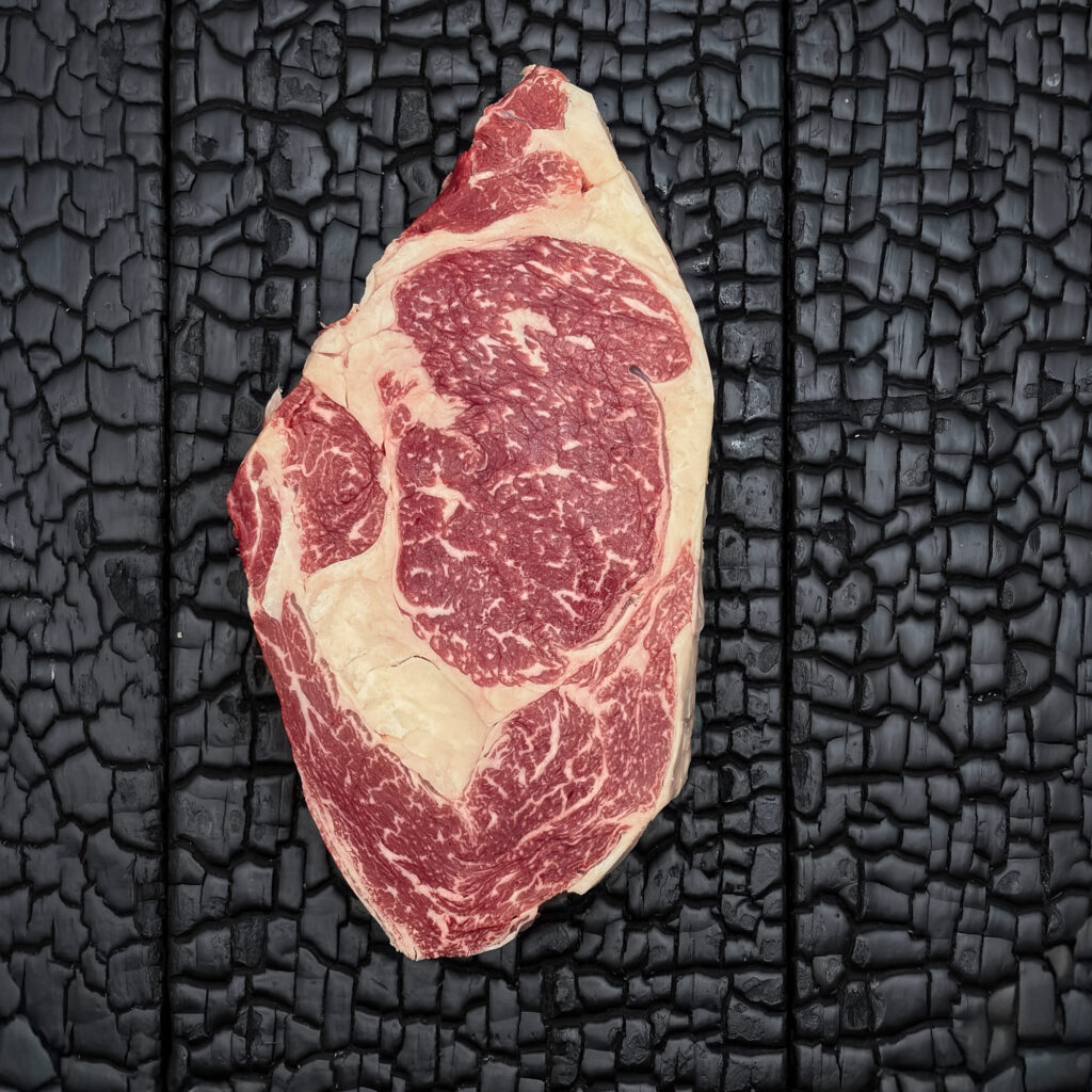 Rohes Rib-Eye-Steak vom Rind mit guter Marmorierung, rote Farbe mit deutlich sichtbarem Fettauge und Fettrand, mittig auf dunkler, verkohlter Holzoberfläche.