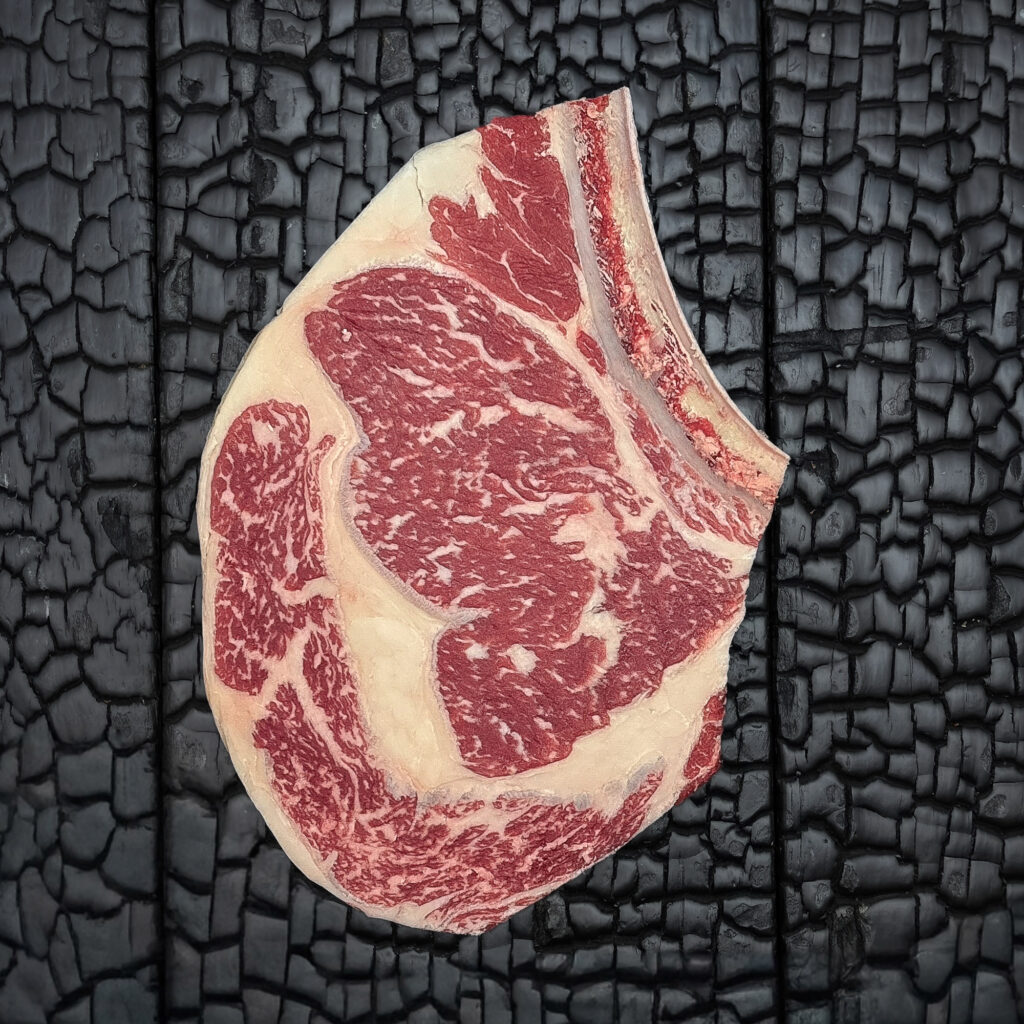 Rohes Rib-Eye-Steak mit Knochen vom Rind, gute Marmorierung, rote Farbe mit sichtbarem Fettauge und Fettrand, mittig auf dunkler, verkohlter Holzoberfläche.