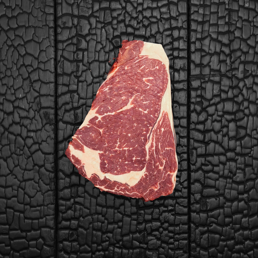 Rohes Rib-Eye-Steak vom Rind mit feiner Marmorierung, rote Farbe mit deutlich sichtbarem Fettauge und Fettrand, mittig auf dunkler, verkohlter Holzoberfläche.
