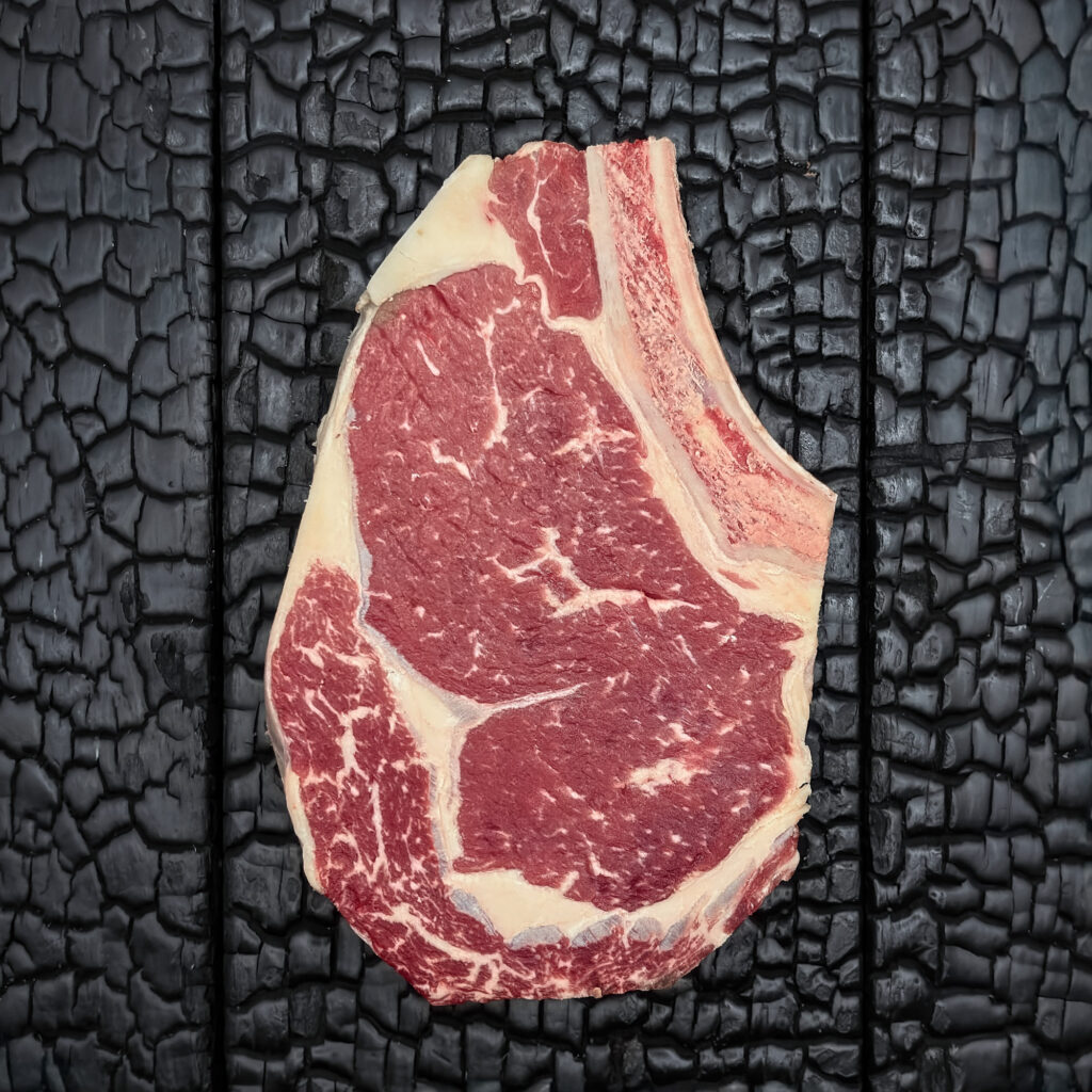 Rohes Rib-Eye-Steak mit Knochen vom Rind, feine Marmorierung, rote Farbe mit sichtbarem Fettauge und Fettrand, mittig auf dunkler, verkohlter Holzoberfläche.