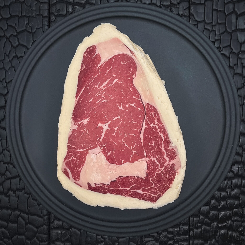 Ein rohes Rib Eye Steak liegt zentral auf einem runden schwarzen Teller. Das dunkelrote Fleisch zeigt eine feine Fettmarmorierung, ein charakteristisches Fettauge und ist von einem hellen Buttermantel umgeben. Der Teller liegt auf einer schwarz verkohlten Holzoberfläche mit stark strukturierter, rissiger Maserung.