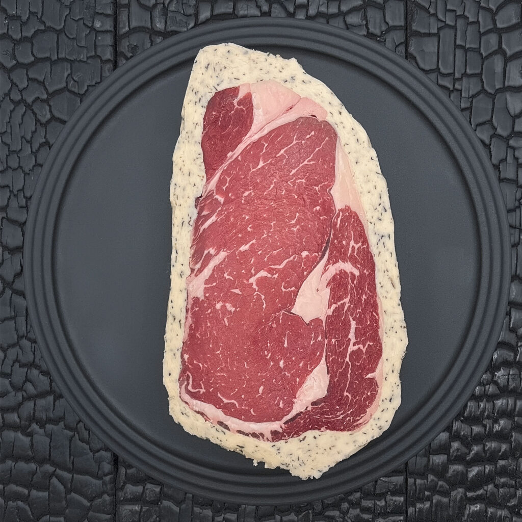 Ein rohes Rib Eye Steak liegt zentral auf einem runden schwarzen Teller. Das dunkelrote Fleisch zeigt eine feine Fettmarmorierung, ein charakteristisches Fettauge und ist von einem hellen Kräuterbuttermantel mit sichtbaren Kräuterstücken umgeben. Der Teller liegt auf einer schwarz verkohlten Holzoberfläche mit stark strukturierter, rissiger Maserung.