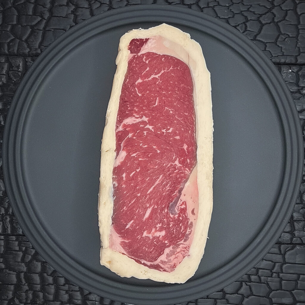 Ein rohes Striploin oder Beiried Steak liegt auf einem runden schwarzen Teller. Das dunkelrote Fleisch zeigt eine feine Fettmarmorierung und ist von einem hellen Buttermantel umgeben, der die gesamte äußere Kontur des Steaks umschließt. Der Teller liegt auf einer schwarz verkohlten Holzoberfläche mit stark strukturierter, rissiger Maserung.