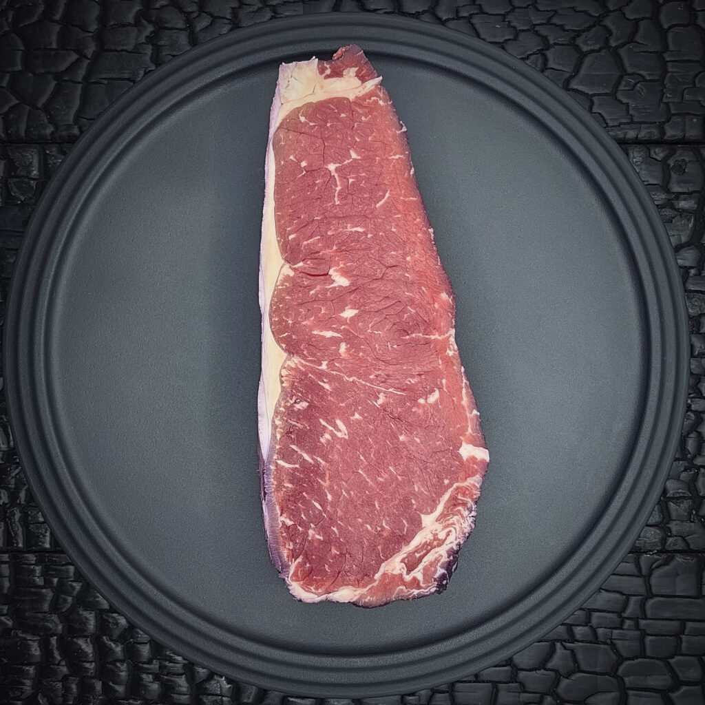 Ein rohes Striploin- oder Beiried-Steak liegt auf einem runden schwarzen Teller. Das rosarote Fleisch zeigt eine feine Fettmarmorierung und einen schmalen hellen Fettrand entlang der Unterseite. Am äußeren Rand sind leicht violette Verfärbungen der Rotweinreifung sichtbar. Die Oberfläche wirkt frisch und gleichmäßig geschnitten. Der Teller liegt auf einer schwarz verkohlten Holzoberfläche mit stark strukturierter, rissiger Maserung.