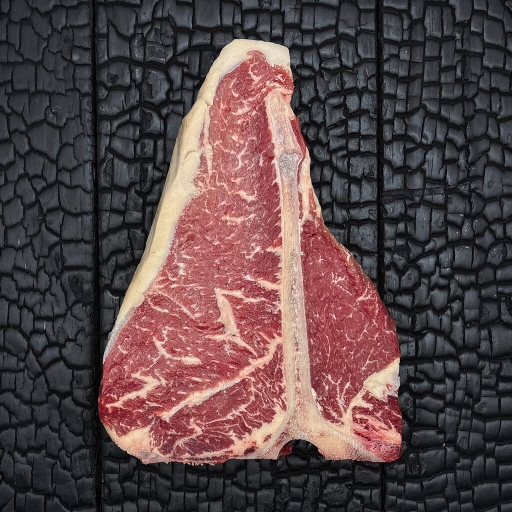 Rohes T-Bone-Steak vom Rind mit T-förmigem Knochen, fein marmoriert, Filet- und Beiriedanteil, rote Farbe mit hellem Fettrand, mittig auf dunkler, verkohlter Holzoberfläche.