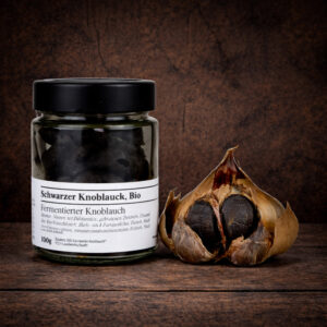 Ein Glas mit schwarzem Schraubdeckel steht links im Bild, gefüllt mit dunklen, fermentierten Knoblauchzehen. Auf dem Etikett ist „Schwarzer Knoblauch, Bio“ sowie „Fermentierter Knoblauch“ zu lesen. Rechts daneben liegt eine aufgebrochene Knoblauchknolle, deren äußere, trockene Schale aufgeklappt ist und mehrere schwarze, weiche Zehen im Inneren freilegt. Die Szene ist mittig auf einer dunklen, rustikalen Holzoberfläche arrangiert.