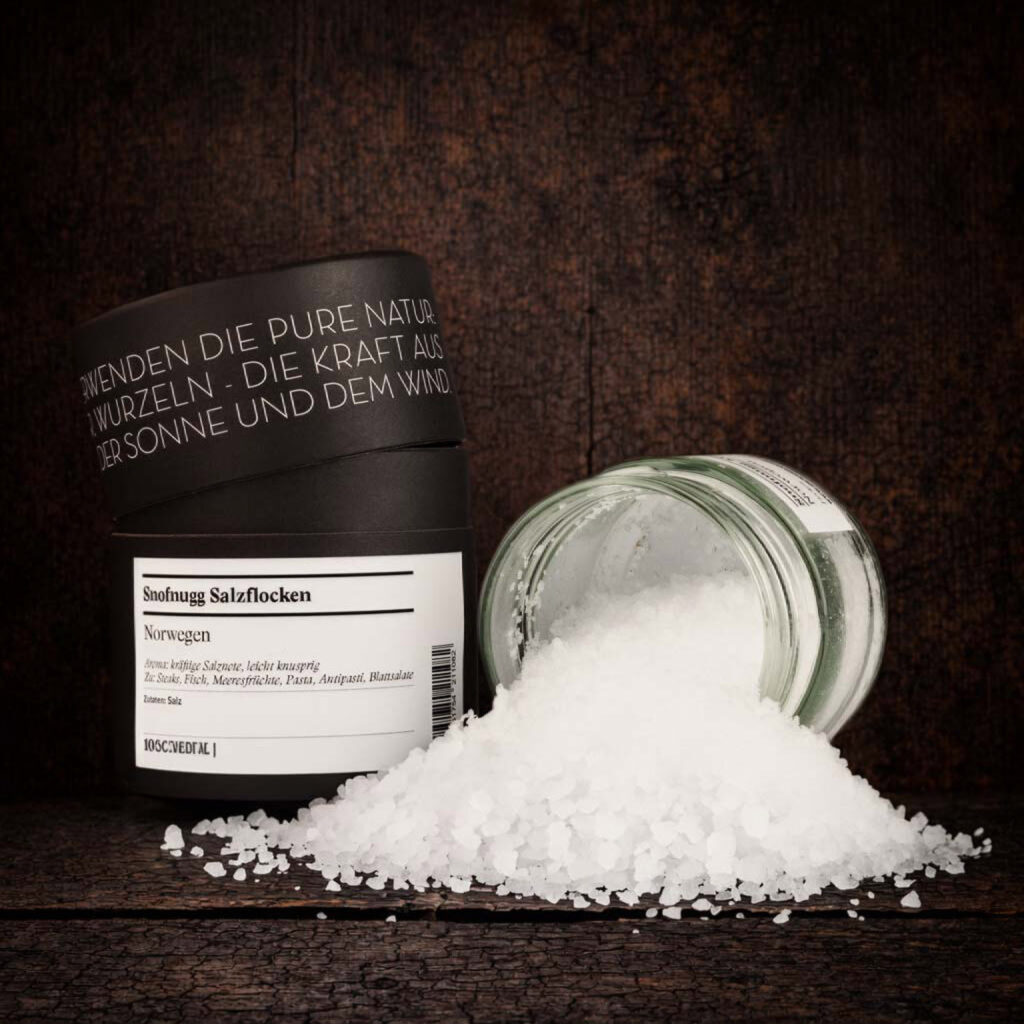 Ein schwarzer Gewürzbehälter mit leicht geöffnetem Deckel steht links im Bild. Auf dem Etikett ist „Snøfnugg Salzflocken“ sowie „Norwegen“ zu lesen. Rechts daneben liegt ein geöffnetes Glas auf der Seite, aus dem grobe, weiße Salzflocken herausgeschüttet wurden und einen kleinen Haufen im Vordergrund bilden. Die Szene ist mittig auf einer dunklen, rustikalen Holzoberfläche arrangiert.