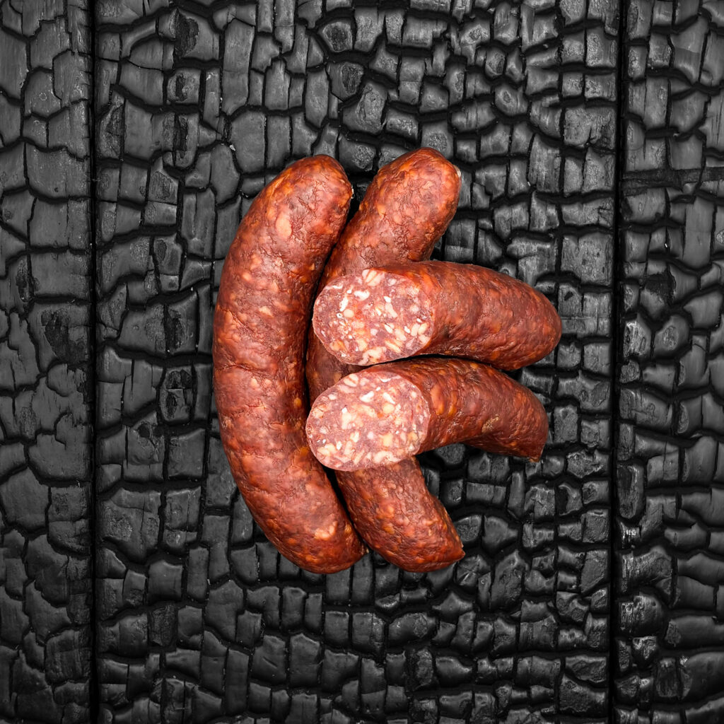 Mehrere Wagyu Jausenwürstel mit Chili und Honig, mit rötlich-brauner, leicht glänzender Wursthülle, locker übereinander angeordnet. Zwei Stück sind quer angeschnitten und zeigen ein grob gekörntes Inneres mit hellen Fettanteilen. Die Würstel liegen mittig auf einer dunklen, verkohlten Holzoberfläche.