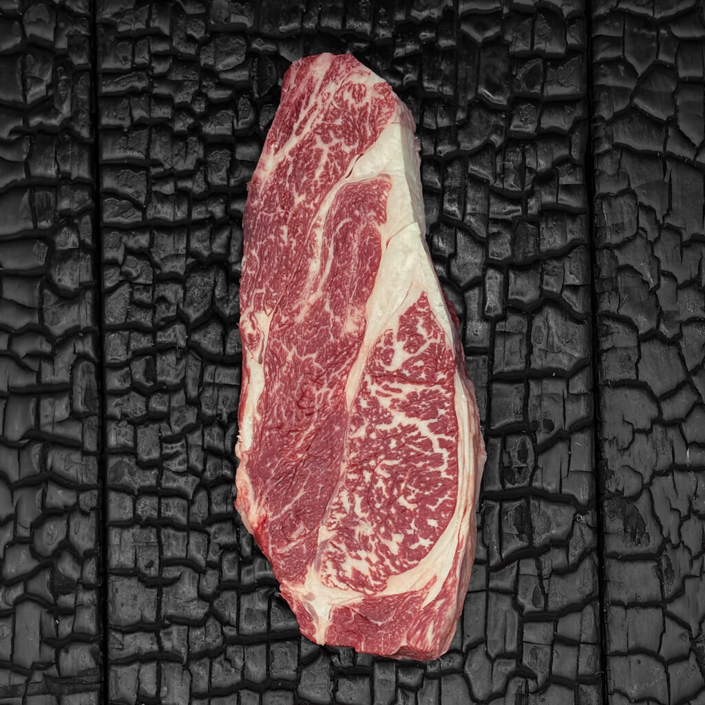 Rohes Chuck-Steak vom Wagyu-Rind, gut marmoriert, rote Fleischfarbe mit sichtbaren Fettadern, mittig auf dunkler, verkohlter Holzoberfläche.