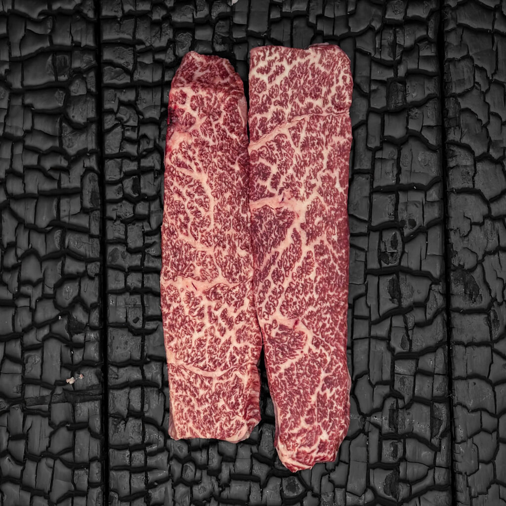 Roher Denver Cut vom Wagyu-Rind, stark marmoriert, in zwei längliche Stücke geschnitten, rote Fleischfarbe, parallel nebeneinander angeordnet, mittig auf dunkler, verkohlter Holzoberfläche.