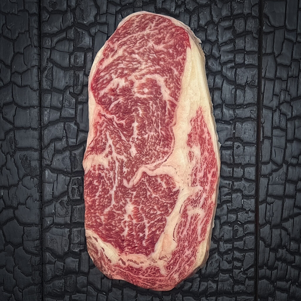 Rohes Entrecote Steak vom Wagyu-Rind, stark marmoriert, rote Fleischfarbe mit hellem Fettrand, mittig auf dunkler, verkohlter Holzoberfläche.