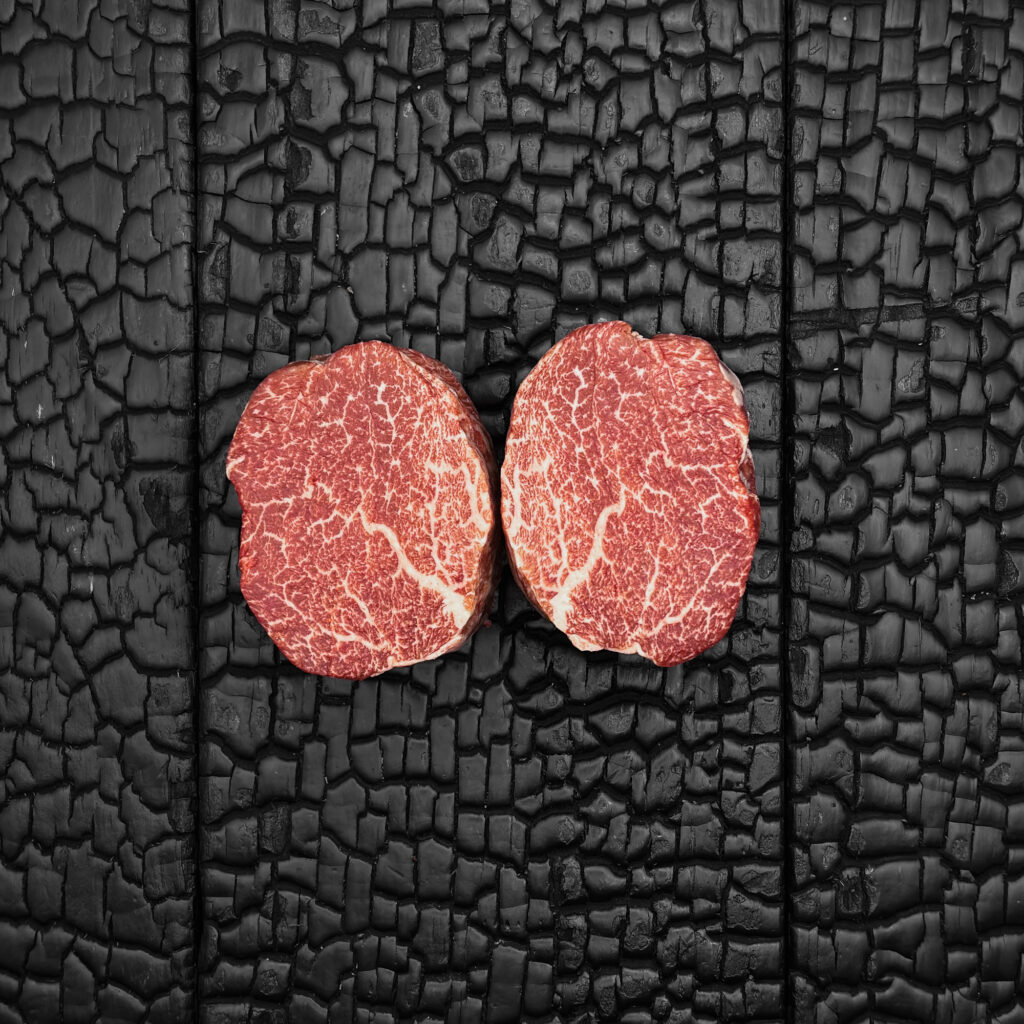 Zwei rohe Filetsteaks vom Wagyu-Rind, stark marmoriert, runde Form, rote Farbe, nebeneinander auf dunkler, verkohlter Holzoberfläche.