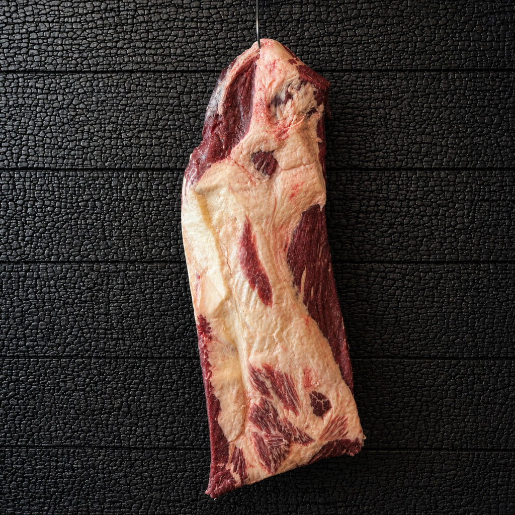 Ein großes längliches Stück rohe Rinderbrust, auch Fullpacker Brisket genannt, hängt vertikal an einem Metallhaken vor einer schwarz verkohlten Holzoberfläche mit stark strukturierter, rissiger Maserung. Das Fleischstück zeigt ausgeprägte helle Fettbereiche sowie dunkelrote Muskelpartien mit sichtbarer Marmorierung und natürlichen Fleischstrukturen. Die Oberfläche wirkt frisch und unbehandelt. Der hohe Fettanteil verteilt sich unregelmäßig über das gesamte Stück und bildet starke Kontraste zum roten Fleisch. Der Metallhaken tritt nur dezent am oberen Bildrand in Erscheinung und unterstreicht die Präsentation im Stil einer Fleischreifung oder traditionellen Metzgerei. Die Aufnahme erfolgt frontal und reduziert. Der dunkle Hintergrund hebt Struktur, Farbe und Form des Fleischstücks deutlich hervor und lenkt den Fokus vollständig auf die Fleischqualität. Die Darstellung vermittelt hochwertige Fleischqualität, traditionelle Fleischreifung und eine handwerkliche Produktinszenierung.