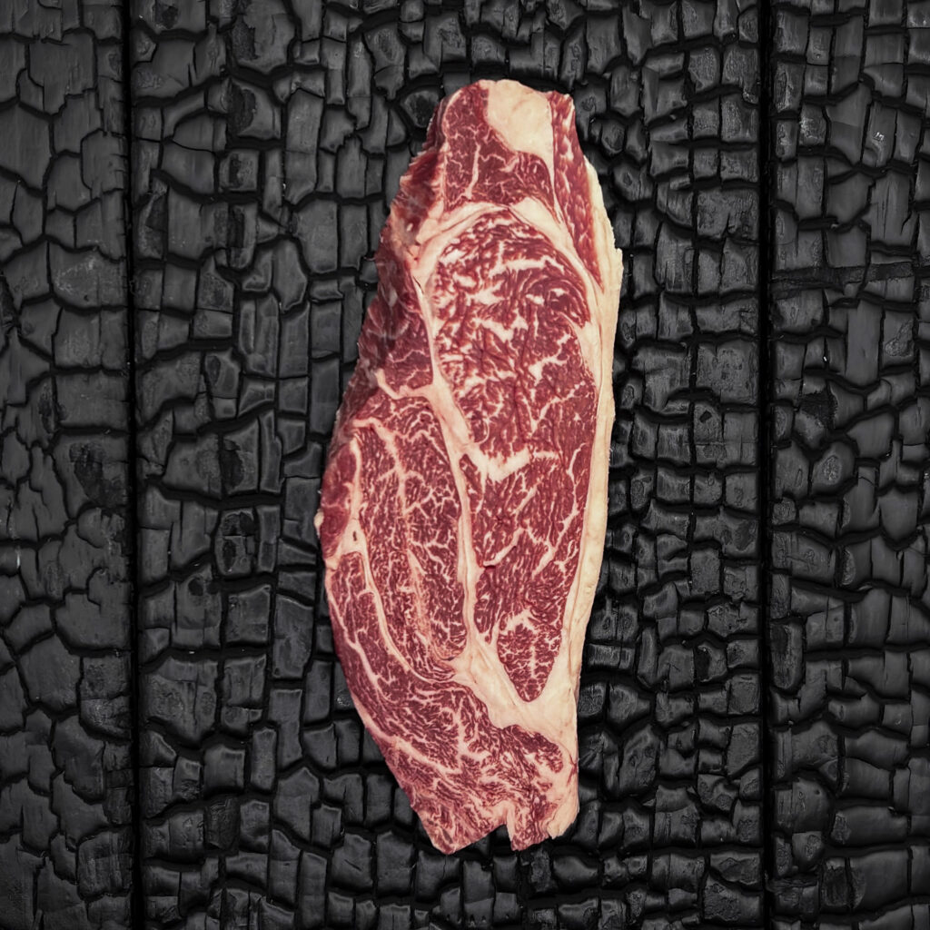 Rohes Chuck-Steak vom Wagyu-Kreuzungsrind, gut marmoriert, rote Farbe, längliche Form, mittig auf dunkler, verkohlter Holzoberfläche.
