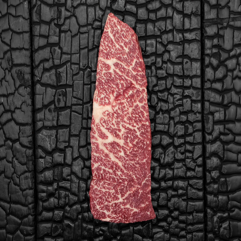 Roher Denver Cut vom Wagyu-Kreuzungsrind, ausgeprägt marmoriert, rote Farbe, längliche Form, mittig auf dunkler, verkohlter Holzoberfläche.