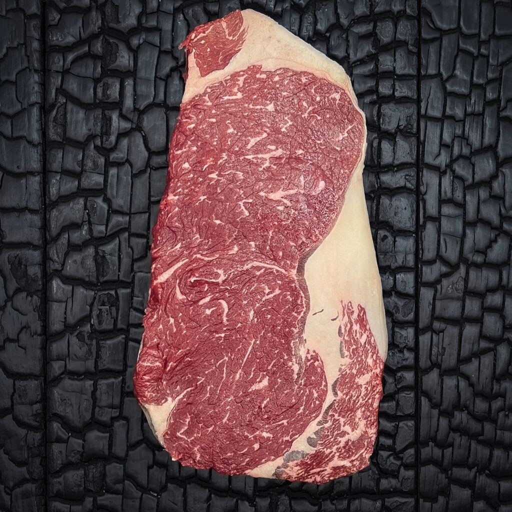 Rohes Entrecôte-Steak vom Wagyu-Kreuzungsrind mit guter Marmorierung, rote Farbe, Fettauge und Fettrand sichtbar, mittig auf dunkler, verkohlter Holzoberfläche.