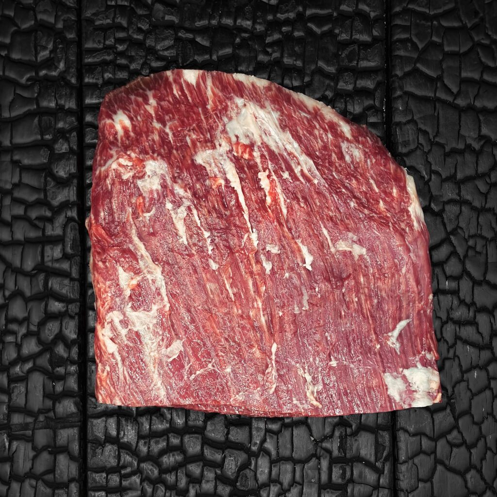Rohes Flank-Steak vom Wagyu-Kreuzungsrind, halbiert, feine Marmorierung, rote Farbe mit sichtbarer Faserstruktur, auf dunkler, verkohlter Holzoberfläche.