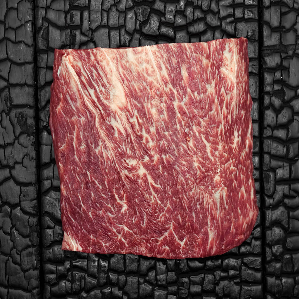 Rohes Flat-Iron-Steak vom Wagyu-Kreuzungsrind, halbiert, gut marmoriert, rote Farbe mit sichtbarer Faserstruktur, auf dunkler, verkohlter Holzoberfläche.