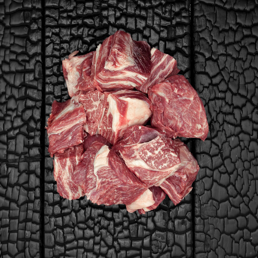 Rohes Gulasch vom Wagyu-Kreuzungsrind, gewürfelt, gut fettdurchzogen, rote Fleischstücke, locker gehäuft, mittig auf dunkler, verkohlter Holzoberfläche.