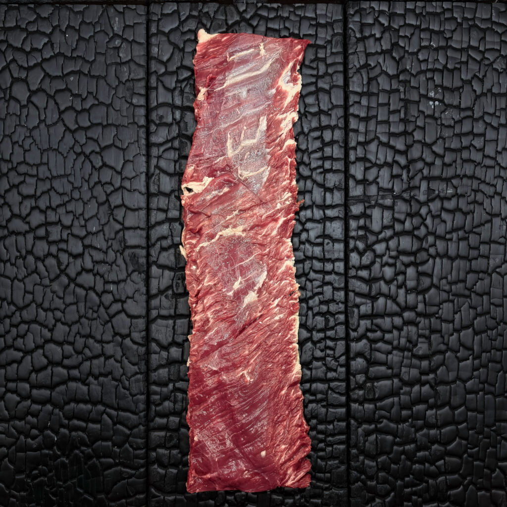 Rohes Outside-Skirt-Steak vom Wagyu-Kreuzungsrind, feine Marmorierung, rote Farbe mit grober Faserstruktur, längliche Form, mittig auf dunkler, verkohlter Holzoberfläche.