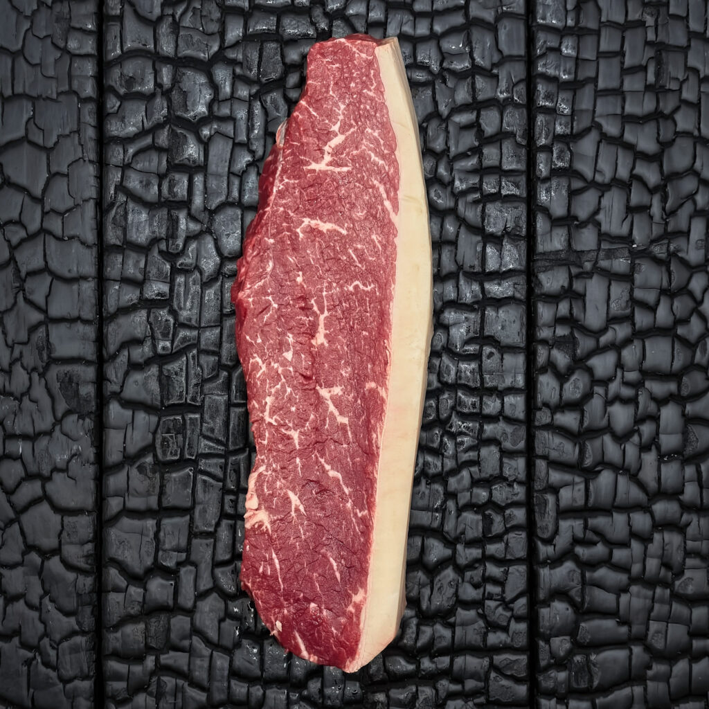 Rohes Picanha-Steak vom Wagyu-Kreuzungsrind, fein marmoriert, rote Farbe mit Fettrand, längliche Form, mittig auf dunkler, verkohlter Holzoberfläche.