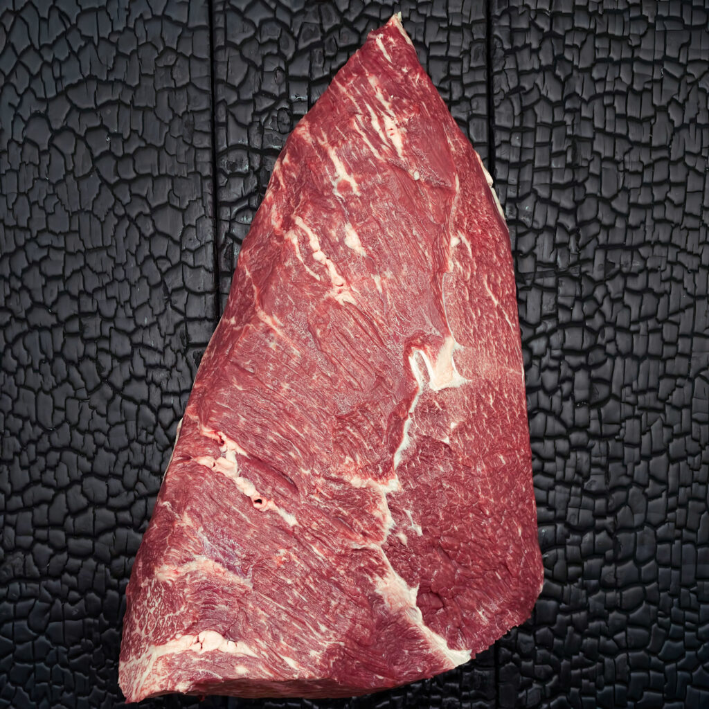Ganzes rohes Picanha vom Wagyu-Kreuzungsrind, dreieckige Form, gut marmoriert, rote Farbe mit Fettdeckel, mittig auf dunkler, verkohlter Holzoberfläche.