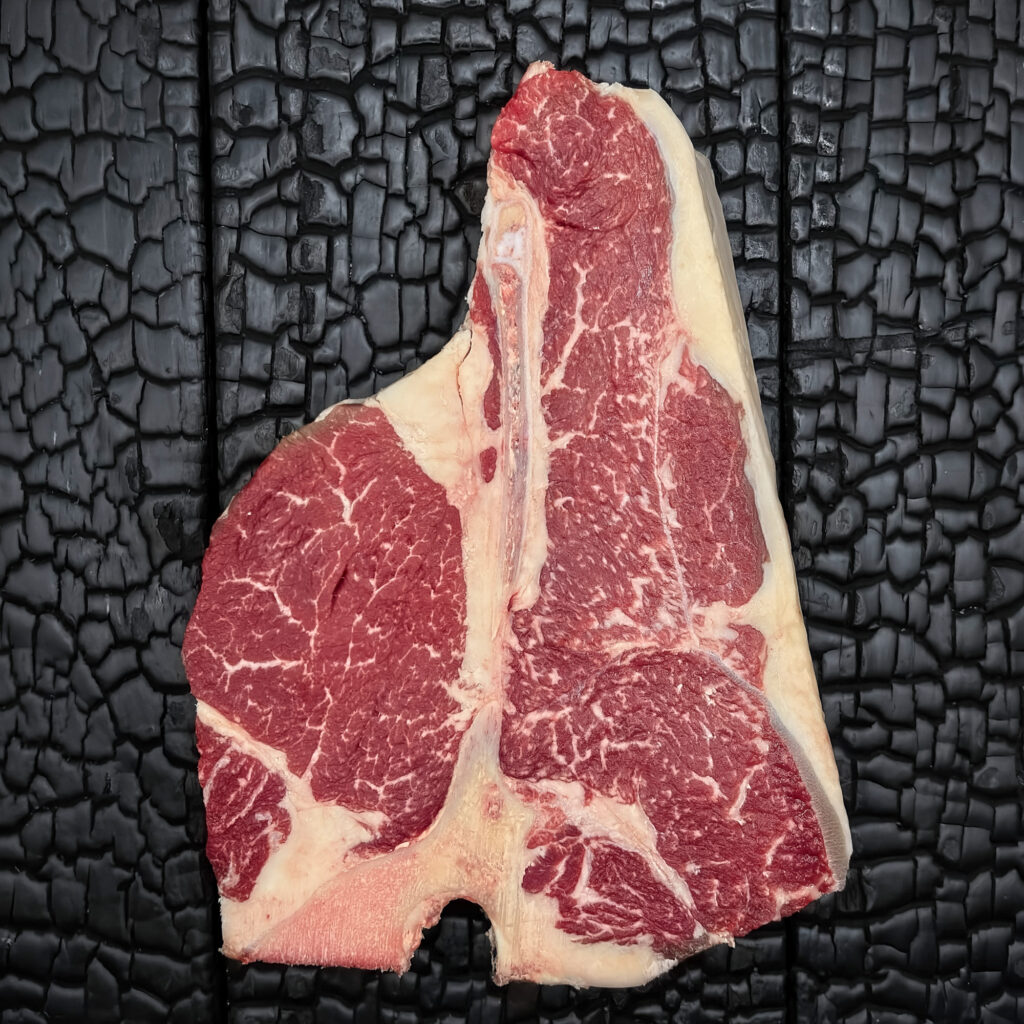 Rohes Porterhouse-Steak mit T-förmigem Knochen vom Wagyu-Kreuzungsrind, gut marmoriert, Filet- und Beiriedanteil, rote Farbe, Fettrand sichtbar, mittig auf dunkler, verkohlter Holzoberfläche.