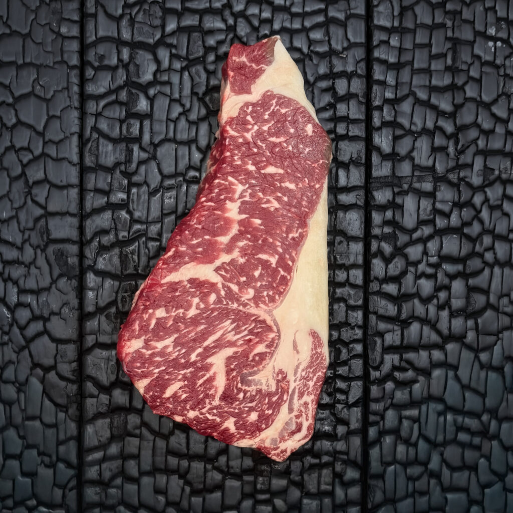 Rohes Entrecôte-Steak vom Wagyu-Kreuzungsrind mit ausgeprägter Marmorierung, rote Farbe, Fettauge und Fettrand sichtbar, mittig auf dunkler, verkohlter Holzoberfläche.