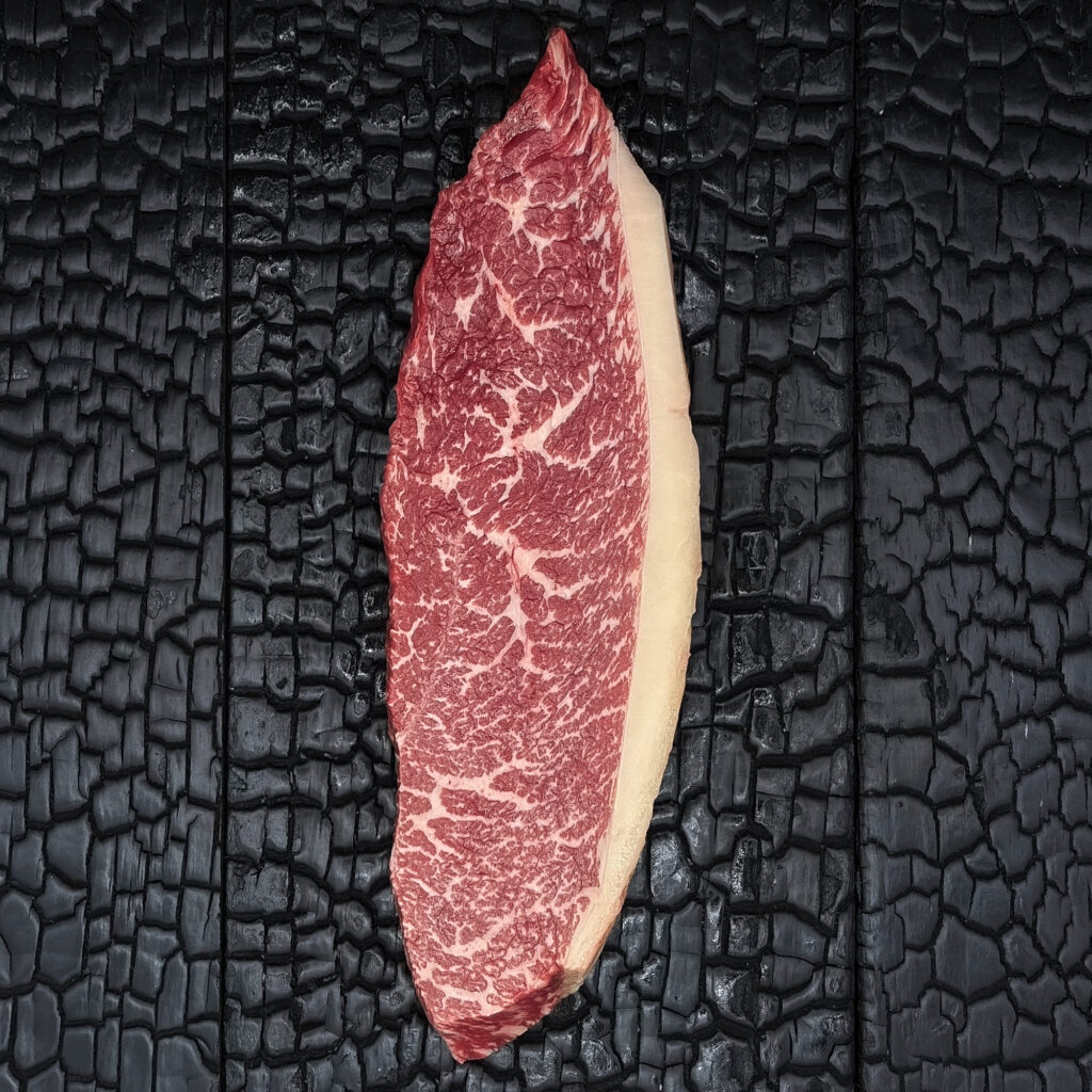 Rohes Picanha-Steak vom Wagyu-Kreuzungsrind, gut marmoriert, rote Farbe mit Fettrand, längliche Form, mittig auf dunkler, verkohlter Holzoberfläche.