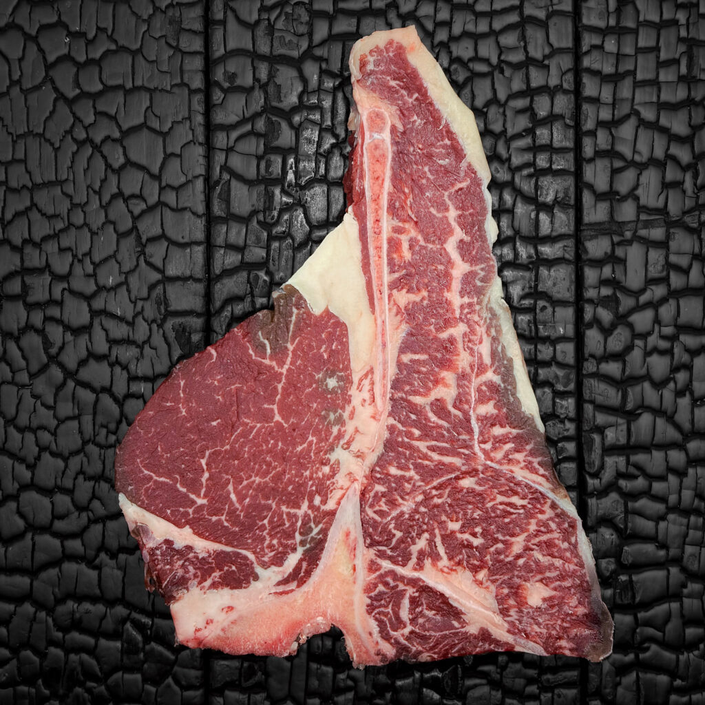 Rohes Porterhouse-Steak mit T-förmigem Knochen vom Wagyu-Kreuzungsrind, ausgeprägt marmoriert, Filet- und Beiriedanteil, rote Farbe, Fettrand sichtbar, mittig auf dunkler, verkohlter Holzoberfläche.