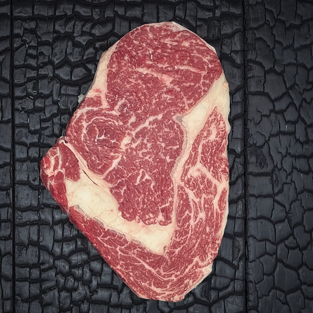 Rohes Rib-Eye-Steak vom Wagyu-Kreuzungsrind mit ausgeprägter Marmorierung, rote Farbe mit deutlich sichtbarem Fettauge und Fettrand, mittig auf dunkler, verkohlter Holzoberfläche.